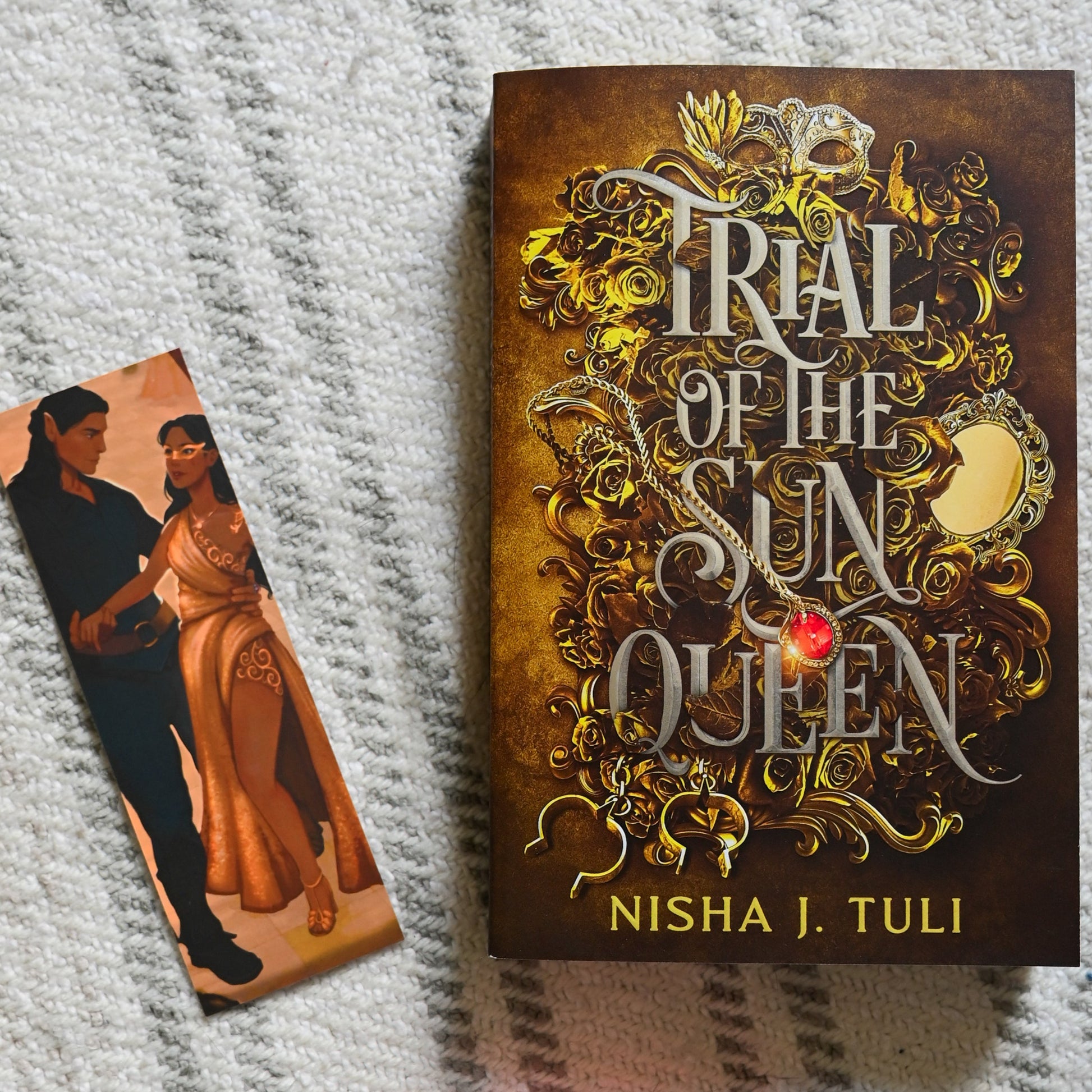 Book titled 'Trial of the Sun Queen' by Nisha J. Tuli on a textured surface with a bookmark and pink flower.
