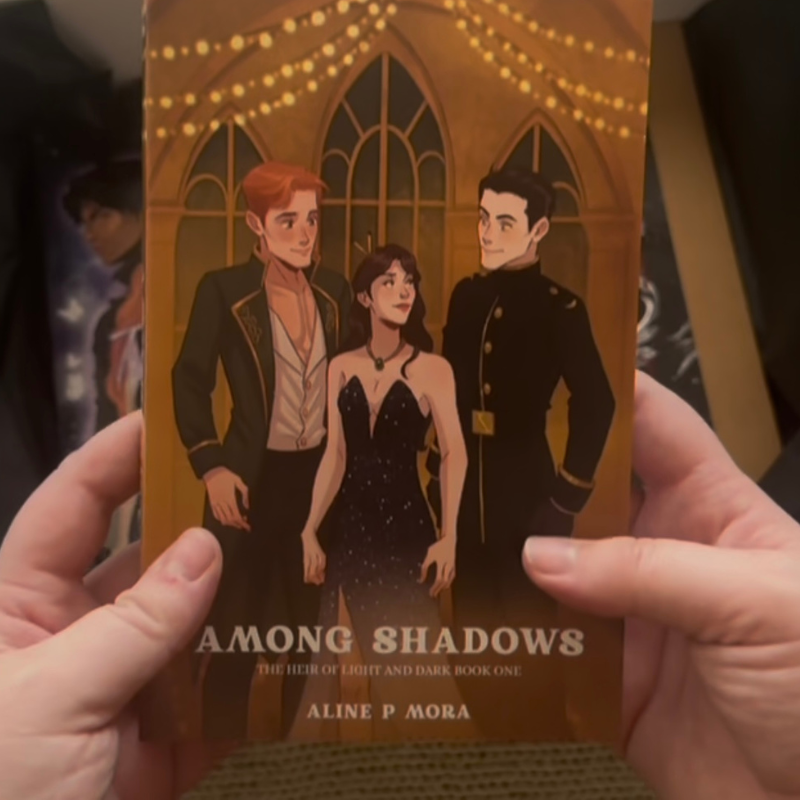 Hands holding Among Shadows by Aline P. Mora — limited signed edition with exclusive Fantasy Bookly character art dust cover featuring three characters under golden lights.